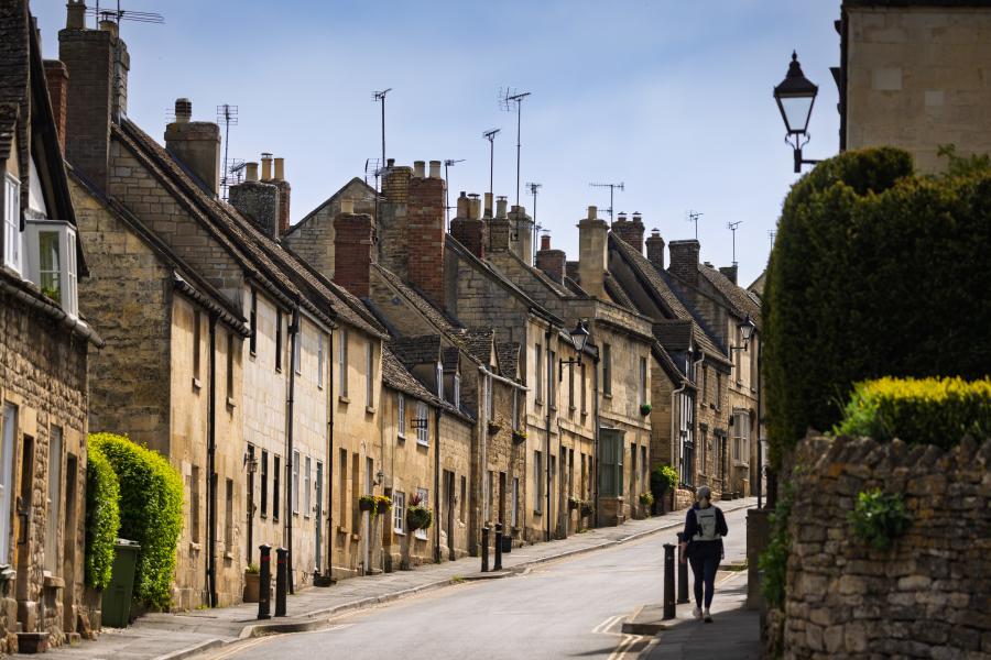 Cotswold Cottage Guesthouse - Wonderful Winchcombe A narrow, gently curving street lined with closely packed stone houses featuring sloped roofs, chimney stacks, and small windows. The buildings vary in warm neutral tones like beige, cream, and light brown, with some greenery climbing walls and hedges along the edges. A single pedestrian walks along the pavement on the right side of the road. A low stone wall and neatly trimmed bush appear in the foreground, while traditional street lamps rise above the homes. The sky overhead is pale and clear, casting soft daylight across the quiet scene in Winchcombe.