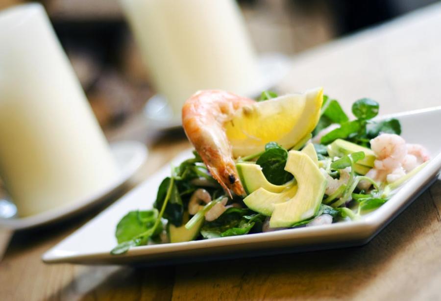 Cotswold Cottage Guesthouse - Wonderful Winchcombe White rectangular plate holding a composed salad with sliced avocado, leafy greens, and pieces of seafood, accompanied by a lemon wedge. The dish is placed on a wooden table, with a softly blurred background featuring a white candle and dining setting, creating a warm restaurant atmosphere.
