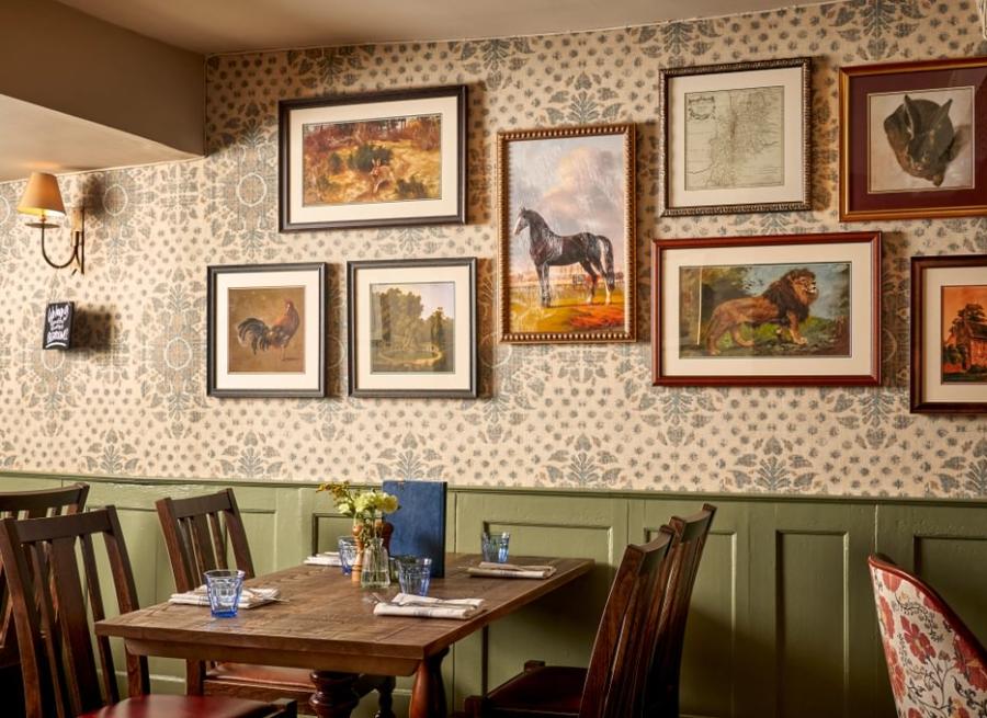 Cotswold Cottage Guesthouse - Wonderful Winchcombe Inside the cosy restaurant at The Lion Inn. A wooden table set for four, surrounded by dark wooden chairs. The wall behind features patterned wallpaper and is decorated with a gallery of framed artwork, many depicting horses and animals in various styles and sizes. A small vase of yellow flowers sits on the table alongside menus, plates, and cutlery. Warm, ambient lighting creates an inviting, traditional atmosphere.